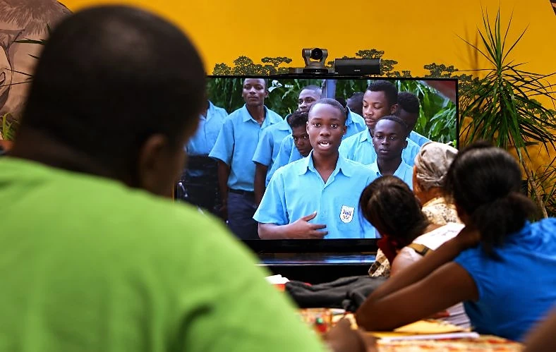 Students watch a large screen as a student in Ghana, in a light-blue school uniform, speaks live with classmates standing behind him
